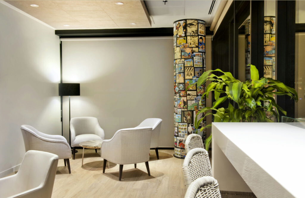 Interiores Minimalistas Acogedores con Sillas de Espuma de Color Gris Claro y Pared de Arte Cómica Un interior minimalista acogedor que presenta tres sillas de espuma de color gris claro alrededor de una pequeña mesa circular, un elegante mostrador blanco con sillas tejidas y una gran planta en maceta, iluminado por una suave luz de una lámpara de pie moderna. La pared trasera exhibe paneles de cómics coloridos, creando un punto focal juguetón en un espacio contemporáneo y relajado.