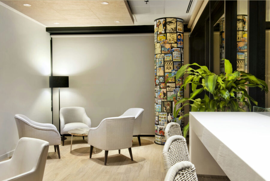 Un interior minimalista acogedor que presenta tres sillas de espuma de color gris claro alrededor de una pequeña mesa circular, un elegante mostrador blanco con sillas tejidas y una gran planta en maceta, iluminado por una suave luz de una lámpara de pie moderna. La pared trasera exhibe paneles de cómics coloridos, creando un punto focal juguetón en un espacio contemporáneo y relajado.