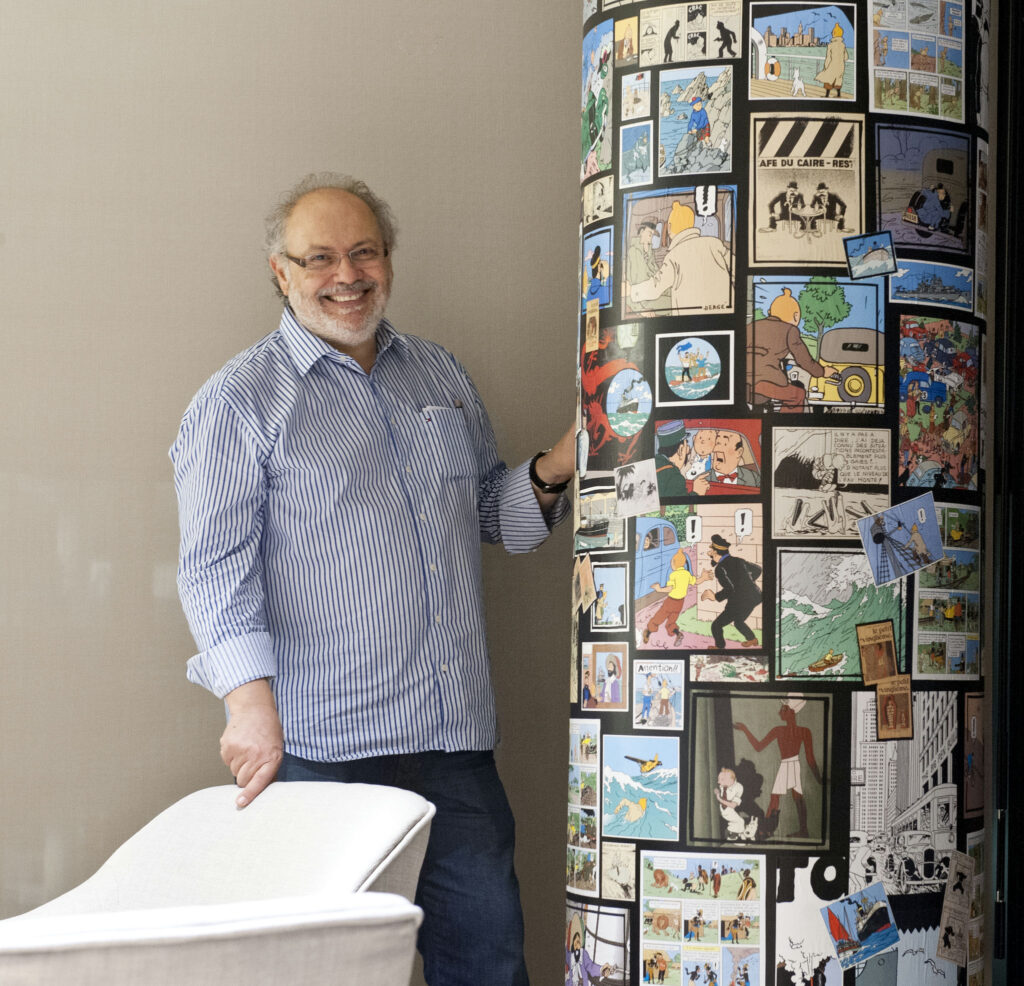 Hombre Alegre de Pie Junto a una Columna Cómica Colorida Un hombre sonriente con cabello rizado y canoso vestido con una camisa de rayas azul y blanca está de pie junto a una alta columna cilíndrica adornada con vibrantes paneles de cómic, que exhiben escenas y personajes caprichosos, contra una pared de color neutro.