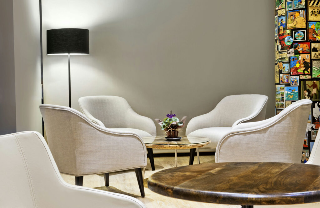 Área de Asientos Moderno y Acogedor con Sillones Blancos y Obras de Arte Coloridas Un acogedor área de asientos que presenta dos sillones blancos tapizados alrededor de una mesa de café de madera, con una pequeña planta en maceta sobre la mesa. Una elegante lámpara de pie negra proporciona una iluminación suave, mientras que una pared vibrante llena de obras de arte eclécticas y coloridas añade un toque animado.