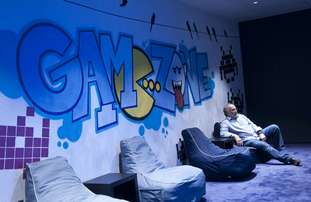 Salón de Juegos Vibrante con Mural Juguetón y Ambiente Relajante Un salón de juegos colorido que presenta un gran mural que dice 'ZONA DE JUEGOS', gráficos juguetones y un personaje de dibujos animados. Un hombre se relaja en un sillón de frijol azul, con una camisa a cuadros azul claro y jeans, mientras una alfombra de color púrpura profundo realza el ambiente divertido.