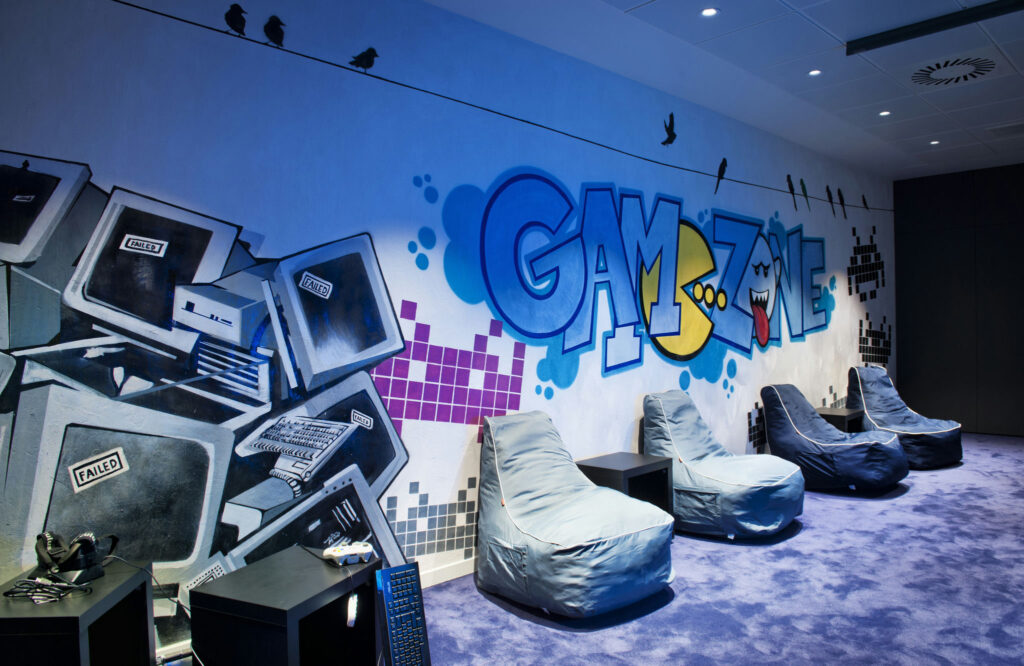 Salón de Juegos Elegante con Mural de Graffiti y Relaxantes Puff Un vibrante salón de juegos que presenta un mural de estilo graffiti que dice 'ZONA DE JUEGOS', gráficos de computadora vintage, cómodos puff, iluminación moderna y suave alfombra púrpura, perfecto para una atmósfera nostálgica de juegos.