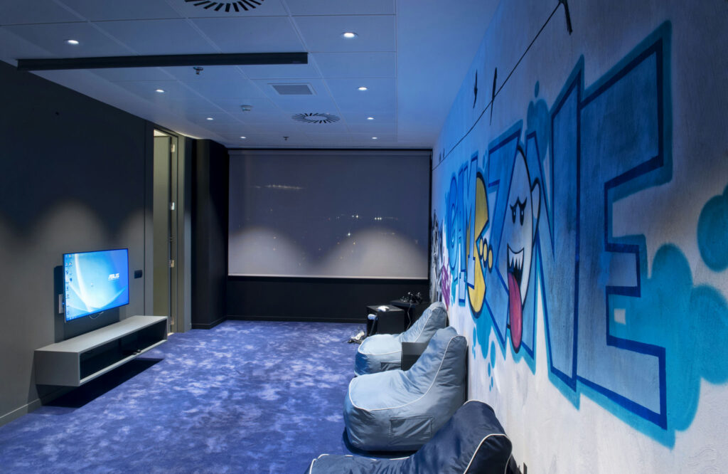 Sala de Juegos Moderna y Acogedora con Toque Artístico Una sala de juegos acogedora y moderna con un televisor de pantalla plana, alfombra azul suave, sillas de frijol azul y grafitis artísticos que dicen 'ZONA DE JUEGO'.