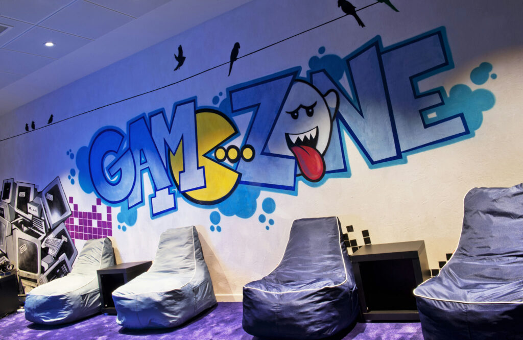 Área de juegos vibrante con mural JUGUETÓN y asientos acogedores Espacio de juegos moderno que exhibe un mural colorido que dice 'ZONA DE JUEGO' con un personaje de fantasma juguetón. El área cuenta con sillas de puff, una pequeña mesa negra y elementos decorativos como computadoras antiguas y arte en píxeles, todo sobre una alfombra morada, creando una atmósfera acogedora para los entusiastas de los videojuegos.