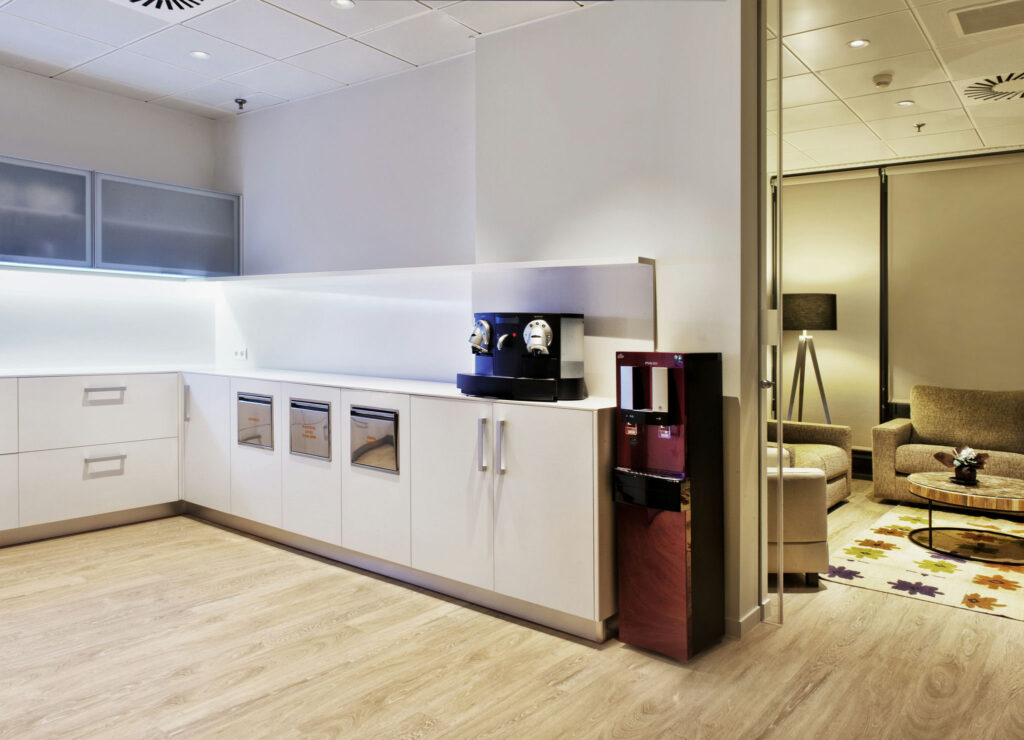 Diseño de Cocina de Oficina Moderna con Área de Salón Acogedora Una cocina de oficina contemporánea que presenta un mobiliario minimalista y elegante, manijas plateadas, una máquina de café centralizada y un dispensador de agua. El espacio está iluminado con luces LED y tiene múltiples cajones. Hay un área de salón elegante en el fondo con un sofá de color marrón claro, una mesa de café redonda y una alfombra con un patrón abstracto.