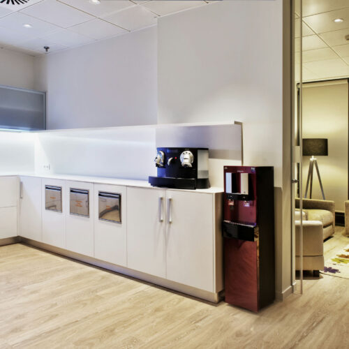 Una cocina de oficina contemporánea que presenta un mobiliario minimalista y elegante, manijas plateadas, una máquina de café centralizada y un dispensador de agua. El espacio está iluminado con luces LED y tiene múltiples cajones. Hay un área de salón elegante en el fondo con un sofá de color marrón claro, una mesa de café redonda y una alfombra con un patrón abstracto.