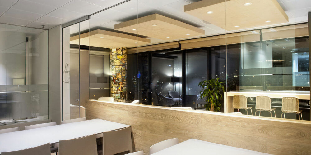 Un interior de oficina contemporáneo con paredes de cristal, una gran mesa blanca, sillas minimalistas y un mostrador de madera con taburetes de bar elegantes, iluminado por una suave iluminación. El fondo muestra obras de arte coloridas y grandes ventanas que ofrecen una vista panorámica del skyline de la ciudad por la noche.