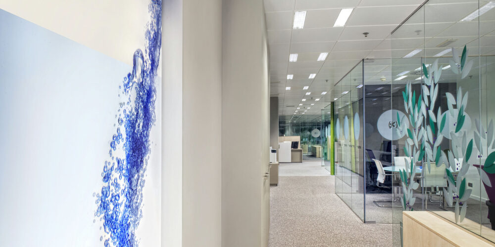 Un interior de oficina contemporáneo que presenta un mural gráfico de agua azul y burbujas, salas de conferencias con paredes de vidrio decoradas con motivos de plantas verdes y muebles de oficina modernos que crean una atmósfera profesional y acogedora.