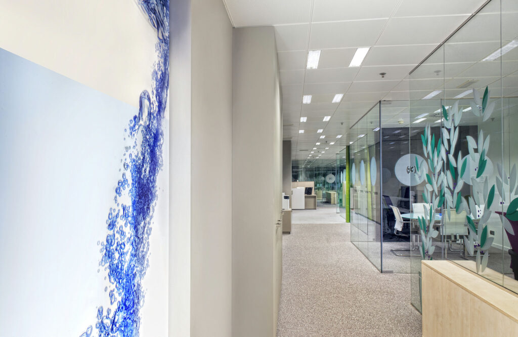 Interior de Oficina Moderna con Diseño Inspirado en la Naturaleza Un interior de oficina contemporáneo que presenta un mural gráfico de agua azul y burbujas, salas de conferencias con paredes de vidrio decoradas con motivos de plantas verdes y muebles de oficina modernos que crean una atmósfera profesional y acogedora.