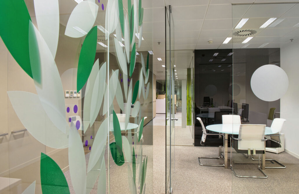 Sala de Reuniones de Oficina Moderna con Paredes de Vidrio Esmerilado y Mobiliario Ergonómico Una sala de reuniones de oficina contemporánea con paredes de vidrio esmerilado decoradas con patrones de hojas verdes y puntos morados. La habitación cuenta con una mesa redonda de vidrio rodeada de sillas ergonómicas blancas, enfatizando un diseño moderno y minimalista. El espacio está bien iluminado y es funcional, con computadoras visibles en el fondo, creando una atmósfera acogedora para la colaboración.