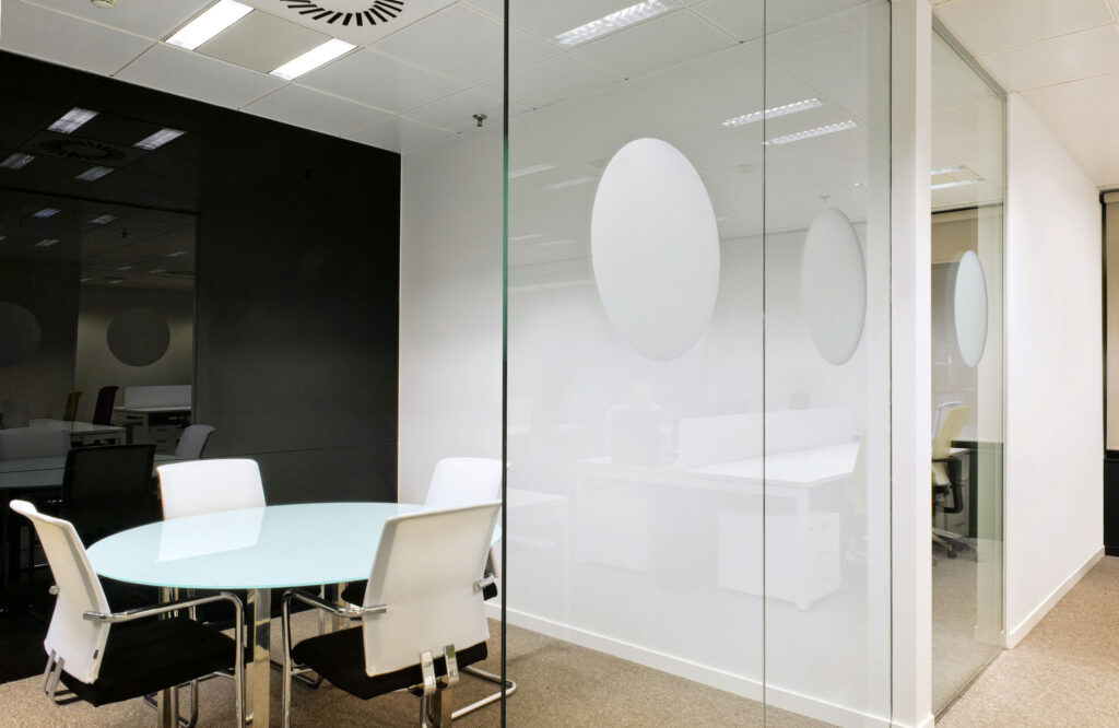 Sala de Reuniones de Oficina Contemporánea con Mesa de Vidrio y Diseño Elegante Una sala de reuniones de oficina moderna que muestra una mesa de vidrio rodeada de sillas elegantes en blanco y negro. La habitación cuenta con una pared de vidrio esmerilado con patrones circulares y está iluminada por luces brillantes en el techo que destacan su estilo contemporáneo.