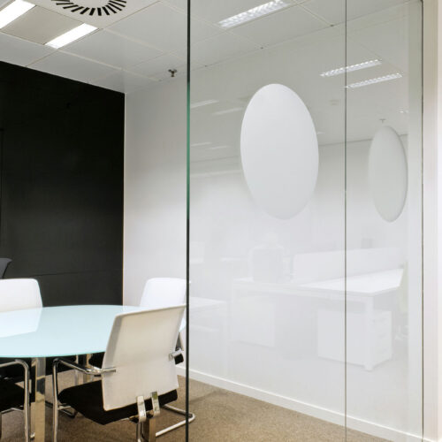 Una sala de reuniones de oficina moderna que muestra una mesa de vidrio rodeada de sillas elegantes en blanco y negro. La habitación cuenta con una pared de vidrio esmerilado con patrones circulares y está iluminada por luces brillantes en el techo que destacan su estilo contemporáneo.
