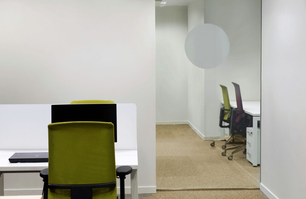 Espacio de Oficina Moderno y Minimalista con Sillas Coloridas Una oficina contemporánea con un diseño minimalista, que presenta una silla de oficina verde en un escritorio blanco elegante con un portátil cerrado. En el fondo, una pared de vidrio revela dos sillas adicionales: una verde y una morada, ubicadas en un ambiente brillante y abierto con paredes de colores claros y una alfombra neutra.