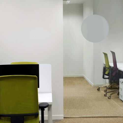 Una oficina contemporánea con un diseño minimalista, que presenta una silla de oficina verde en un escritorio blanco elegante con un portátil cerrado. En el fondo, una pared de vidrio revela dos sillas adicionales: una verde y una morada, ubicadas en un ambiente brillante y abierto con paredes de colores claros y una alfombra neutra.