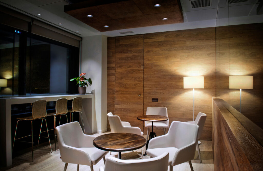 Lounge Moderno Acogedor con Vista a la Ciudad y Decoración Elegante Un área de lounge estilosa con sillas tapizadas blancas alrededor de mesas de madera, una lámpara de pie cálida, una planta decorativa en maceta y un bar elegante con taburetes en tonos dorados que muestra una vista impresionante del paisaje urbano nocturno a través de grandes ventanas, complementada por ricas paredes de madera.