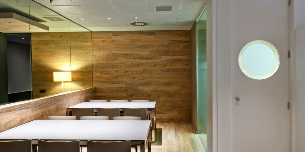 Una sala de reuniones moderna y elegante que presenta una larga mesa blanca rodeada de sillas beige. El espacio se realza con una pared de acento de madera y una suave iluminación ambiental de una lámpara de pared. Una ventana redonda en el fondo ofrece una vista de otra área, mientras que un gran espejo refleja la amplitud de la sala, y el suelo de madera clara completa el aspecto contemporáneo.