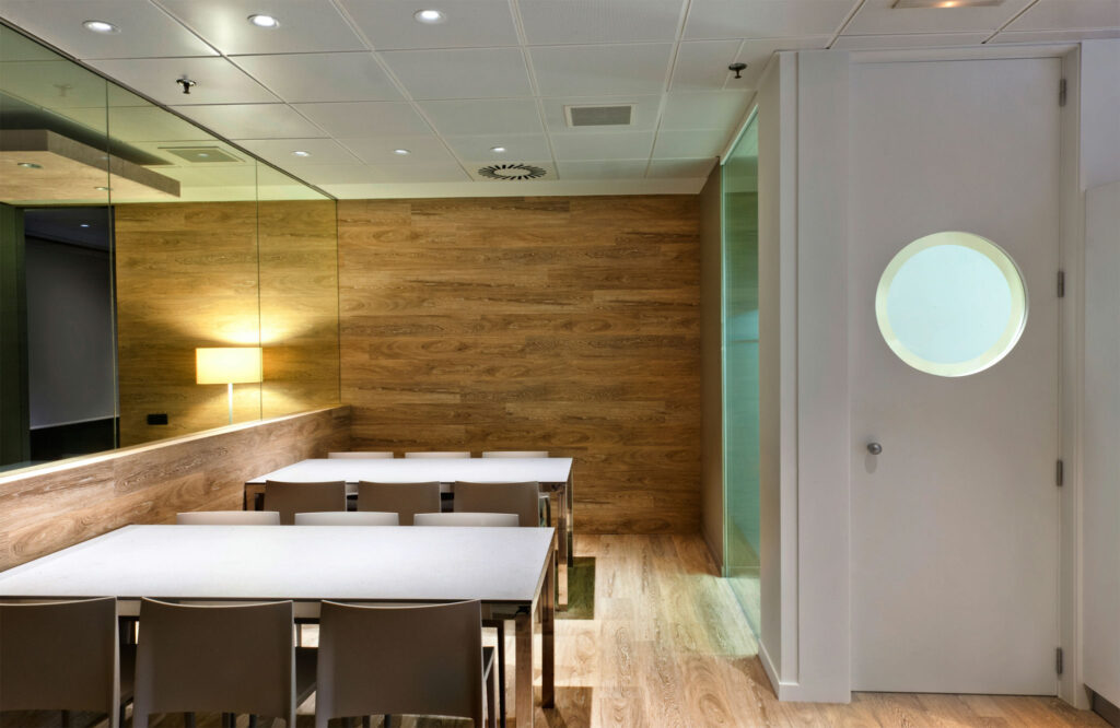 Sala de Conferencias Moderna con Acentos de Madera y Iluminación Cálida Una sala de reuniones moderna y elegante que presenta una larga mesa blanca rodeada de sillas beige. El espacio se realza con una pared de acento de madera y una suave iluminación ambiental de una lámpara de pared. Una ventana redonda en el fondo ofrece una vista de otra área, mientras que un gran espejo refleja la amplitud de la sala, y el suelo de madera clara completa el aspecto contemporáneo.