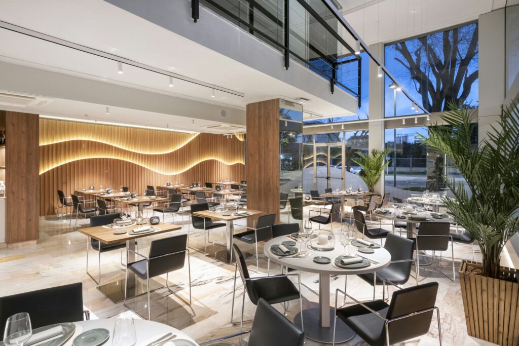 Restaurante Moderno con Diseño Interior Elegante Diseño interior contemporáneo de un restaurante con mesas de madera, sillas negras, y paredes de madera iluminadas a rayas. Espacio luminoso con grandes ventanales y plantas decorativas.