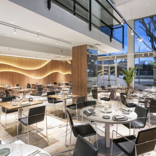 Diseño interior contemporáneo de un restaurante con mesas de madera, sillas negras, y paredes de madera iluminadas a rayas. Espacio luminoso con grandes ventanales y plantas decorativas.