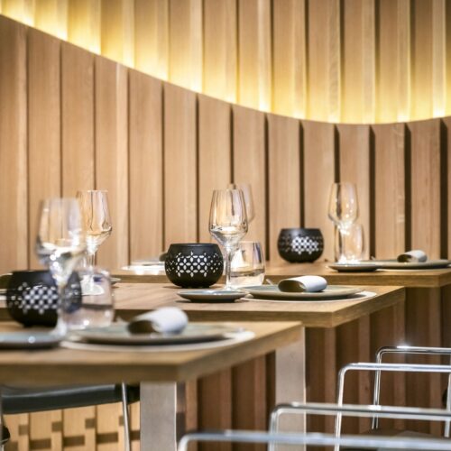 Comedor elegante con mesas de madera, sillas de acrílico transparente y vajilla minimalista en tonos negros y dorados.