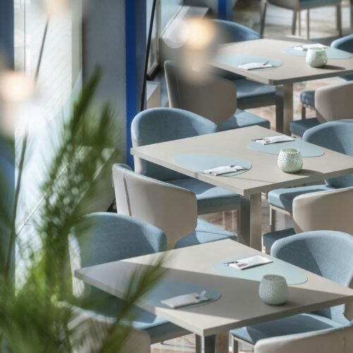 Interior de un restaurante moderno con mesas de madera clara, sillas tapizadas en azul y decoración minimalista que integra elementos naturales.