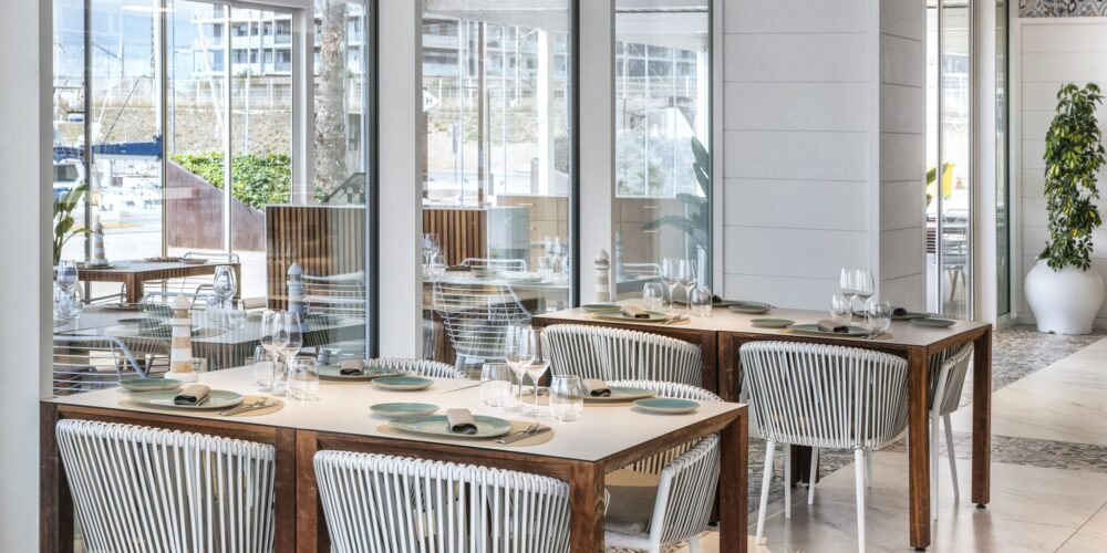 Vista interior de un restaurante elegante y moderno que presenta mesas de madera emparejadas con sillas de mimbre blancas. El amplio comedor está iluminado por luz natural que entra a través de grandes ventanales de vidrio, revelando la vegetación exterior. Las mesas están decoradas con elegantes platos y cubiertos, lo que contribuye a la atmósfera acogedora creada por la estética limpia y la decoración contemporánea.