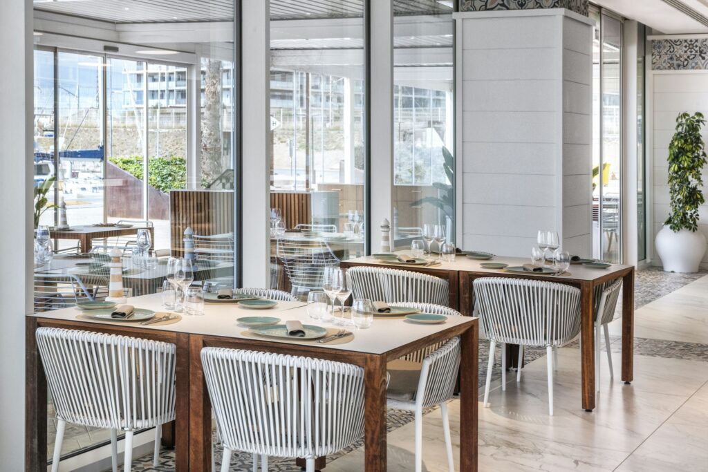 Moderno interior de restaurante luminoso y espacioso con mesas de madera y sillas de mimbre Vista interior de un restaurante elegante y moderno que presenta mesas de madera emparejadas con sillas de mimbre blancas. El amplio comedor está iluminado por luz natural que entra a través de grandes ventanales de vidrio, revelando la vegetación exterior. Las mesas están decoradas con elegantes platos y cubiertos, lo que contribuye a la atmósfera acogedora creada por la estética limpia y la decoración contemporánea.