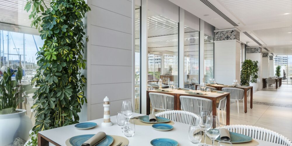 Una mesa de comedor elegantemente decorada en un restaurante moderno, con platos azules, servilletas plateadas y copas de vino. Plantas verdes exuberantes y grandes ventanales de cristal que muestran vistas al puerto crean una atmósfera acogedora.