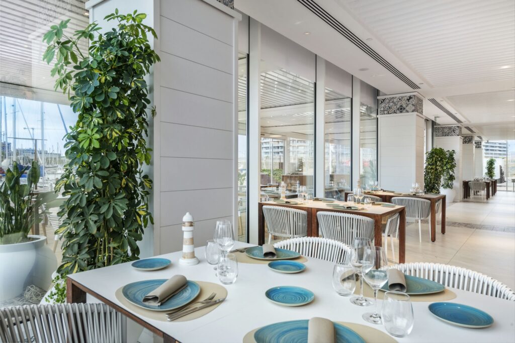 Áreas de Comedor Moderno y Elegante con Vistas al Puerto Una mesa de comedor elegantemente decorada en un restaurante moderno, con platos azules, servilletas plateadas y copas de vino. Plantas verdes exuberantes y grandes ventanales de cristal que muestran vistas al puerto crean una atmósfera acogedora.