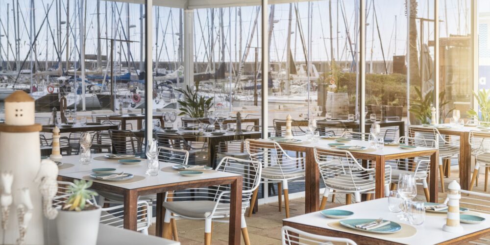 Un interior de restaurante moderno con mesas de comedor dispuestas ordenadamente y decoradas con elegante vajilla y cristalería. Los amplios paneles de vidrio ofrecen una vista panorámica de una marina llena de barcos y veleros. El espacio luminoso y aireado cuenta con mobiliario blanco y de madera estiloso, complementado con plantas en macetas que añaden verdor, creando una atmósfera de comedor tranquila, perfecta para disfrutar de comidas junto al agua.