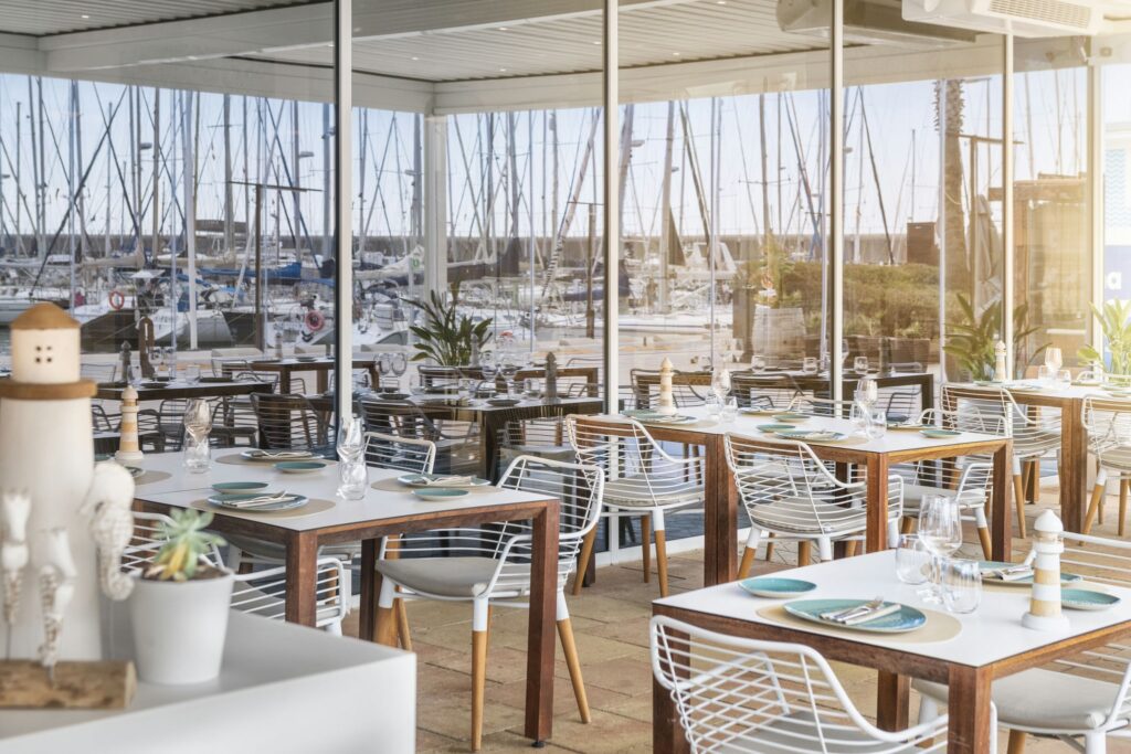 Interior Elegante de Restaurante Costero con Vista a la Marina Un interior de restaurante moderno con mesas de comedor dispuestas ordenadamente y decoradas con elegante vajilla y cristalería. Los amplios paneles de vidrio ofrecen una vista panorámica de una marina llena de barcos y veleros. El espacio luminoso y aireado cuenta con mobiliario blanco y de madera estiloso, complementado con plantas en macetas que añaden verdor, creando una atmósfera de comedor tranquila, perfecta para disfrutar de comidas junto al agua.