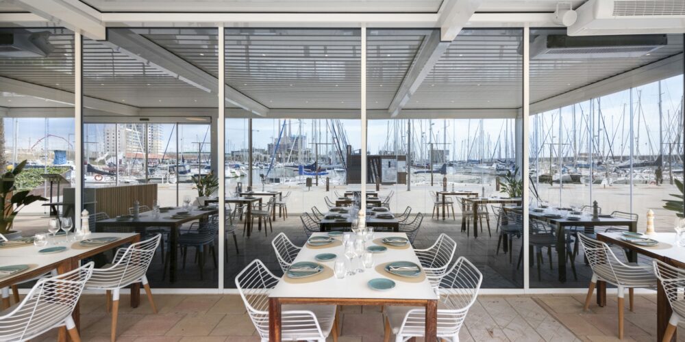 Un interior contemporáneo de restaurante con grandes ventanales de vidrio que muestran un pintoresco puerto lleno de veleros. El área de comedor incluye mesas de madera elegantes adornadas con platos y cristalería sofisticada, acompañadas de sillas de alambre blanco que realzan la sensación de ligereza y amplitud del espacio. La luz del sol entra a raudales, iluminando los elementos de diseño elegantes y la atmósfera acogedora, mientras que el área de asientos al aire libre añade un encanto náutico.