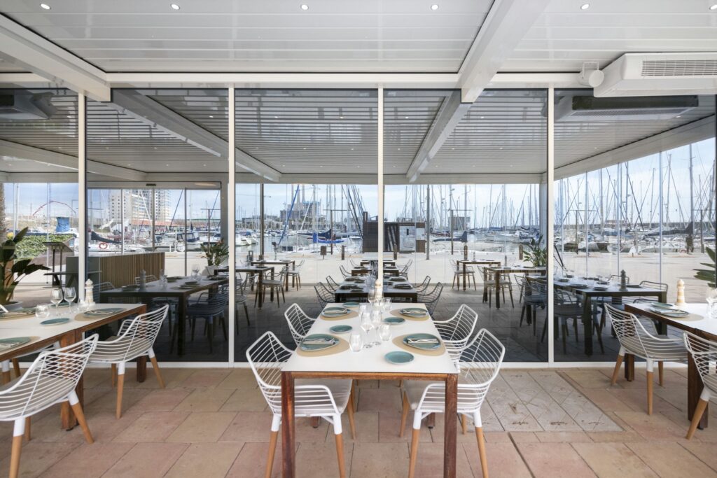 Restaurante Moderno Chic con Vista al Puerto Un interior contemporáneo de restaurante con grandes ventanales de vidrio que muestran un pintoresco puerto lleno de veleros. El área de comedor incluye mesas de madera elegantes adornadas con platos y cristalería sofisticada, acompañadas de sillas de alambre blanco que realzan la sensación de ligereza y amplitud del espacio. La luz del sol entra a raudales, iluminando los elementos de diseño elegantes y la atmósfera acogedora, mientras que el área de asientos al aire libre añade un encanto náutico.