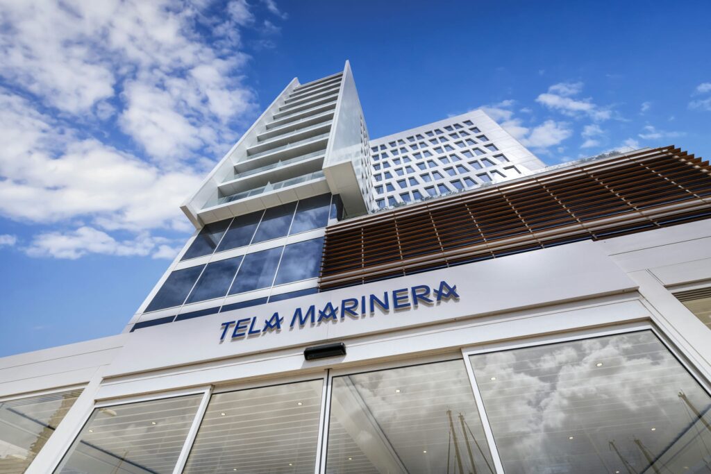 TELA MARINERA: Una Maravilla Arquitectónica Moderna Un edificio contemporáneo y elegante llamado 'TELA MARINERA' con grandes ventanales de vidrio y una fachada de varios niveles, enmarcado por un cielo azul brillante con nubes blancas esponjosas.