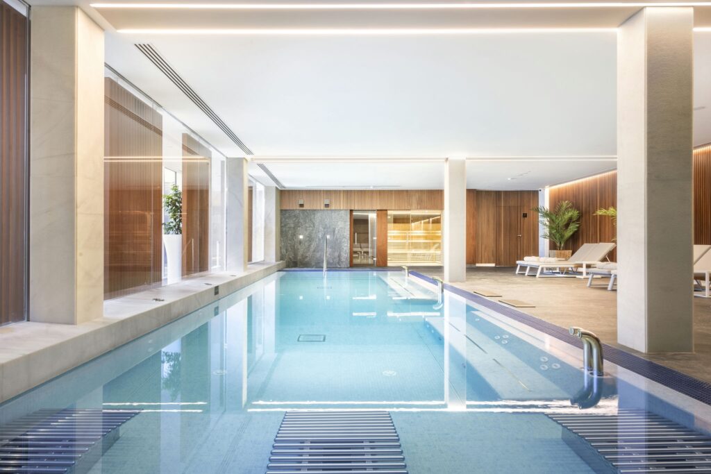Moderna piscina interior con diseño elegante y área de relajación Una piscina interior contemporánea con una superficie limpia de agua azul, rodeada de grandes ventanas y paneles de madera elegantes, iluminada por suaves luces de techo. Cerca hay tumbonas para relajación y una planta en maceta verde, creando una atmósfera tranquila e invitante.