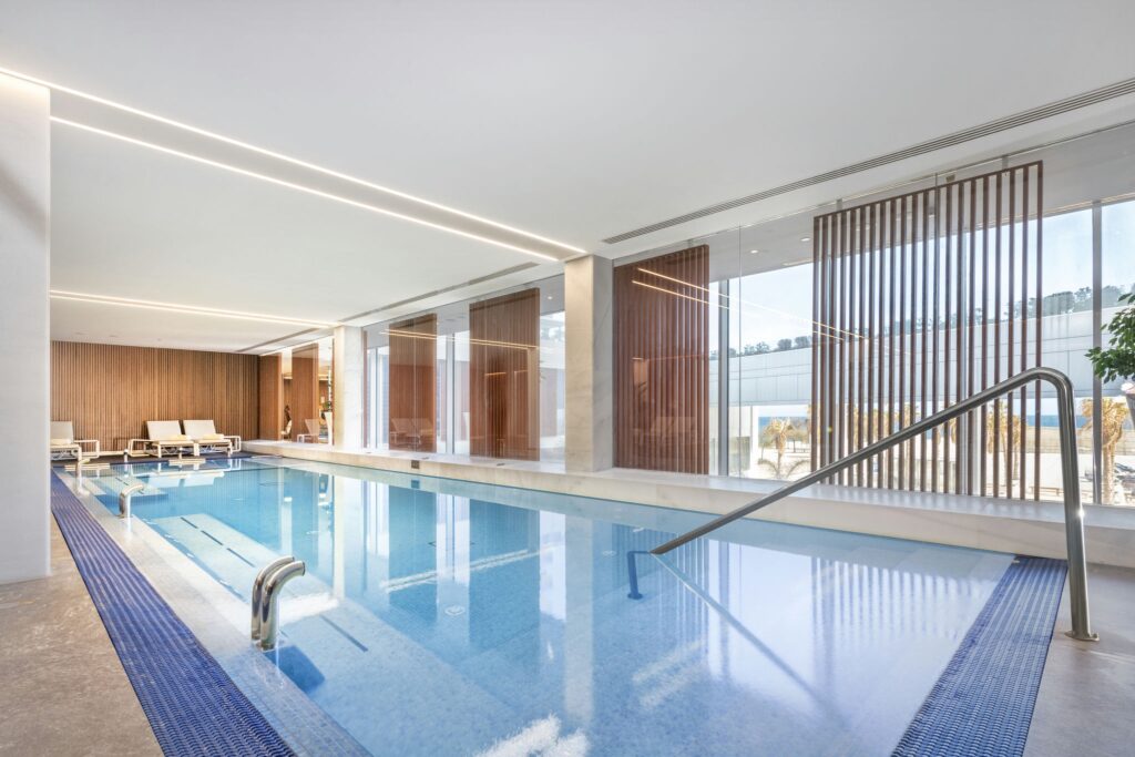 Moderna piscina cubierta con diseño minimalista y luz natural Una serena piscina cubierta que muestra una característica de agua azul clara, rodeada de grandes ventanas de vidrio que iluminan el espacio con luz natural. El diseño minimalista se complementa con acentos de madera clara y sillas de lounge contemporáneas, creando una atmósfera acogedora.