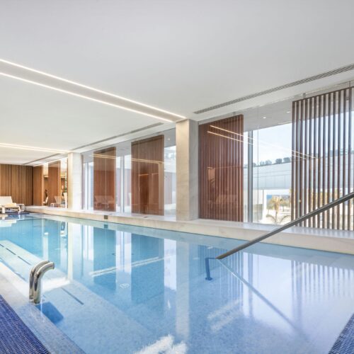 Una serena piscina cubierta que muestra una característica de agua azul clara, rodeada de grandes ventanas de vidrio que iluminan el espacio con luz natural. El diseño minimalista se complementa con acentos de madera clara y sillas de lounge contemporáneas, creando una atmósfera acogedora.