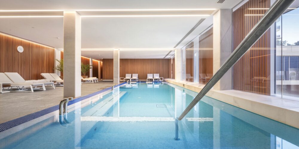 Una elegante piscina interior que presenta agua azul clara, modernas sillas de descanso y elementos de diseño en madera. El espacio cuenta con abundante luz natural proveniente de grandes ventanas, creando una atmósfera serena complementada por una iluminación cálida que realza las características arquitectónicas modernas.