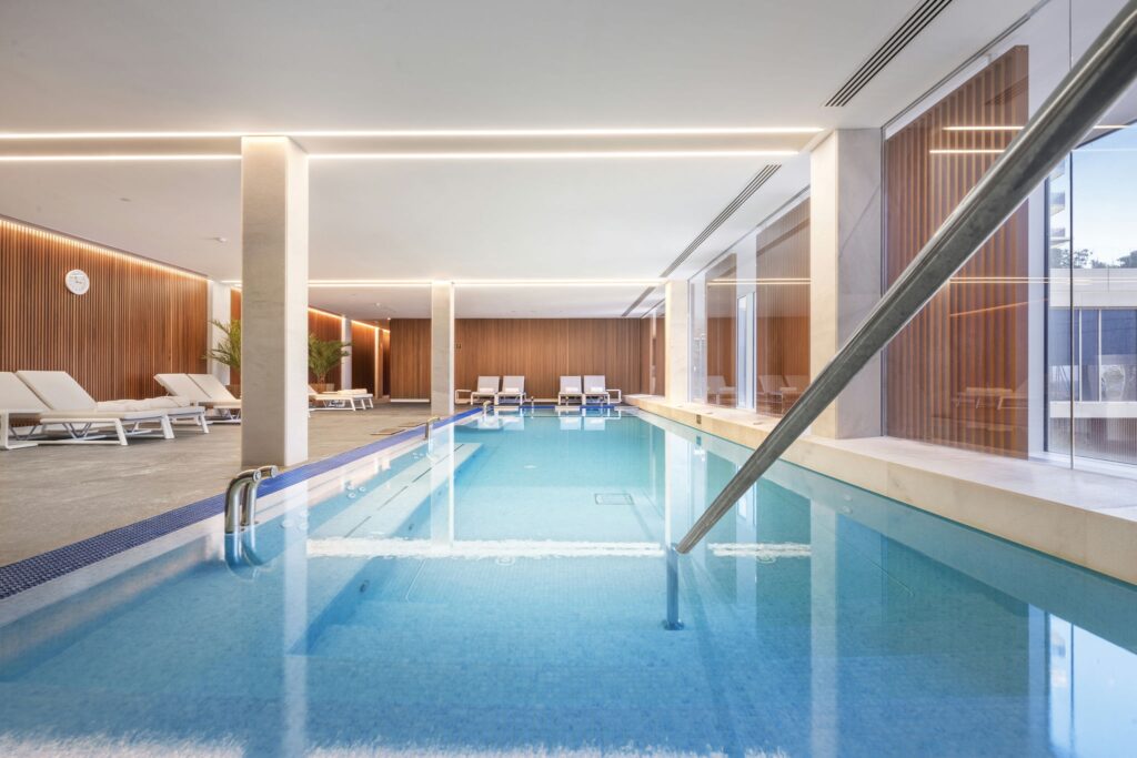 Elegante piscina interior con arquitectura moderna Una elegante piscina interior que presenta agua azul clara, modernas sillas de descanso y elementos de diseño en madera. El espacio cuenta con abundante luz natural proveniente de grandes ventanas, creando una atmósfera serena complementada por una iluminación cálida que realza las características arquitectónicas modernas.