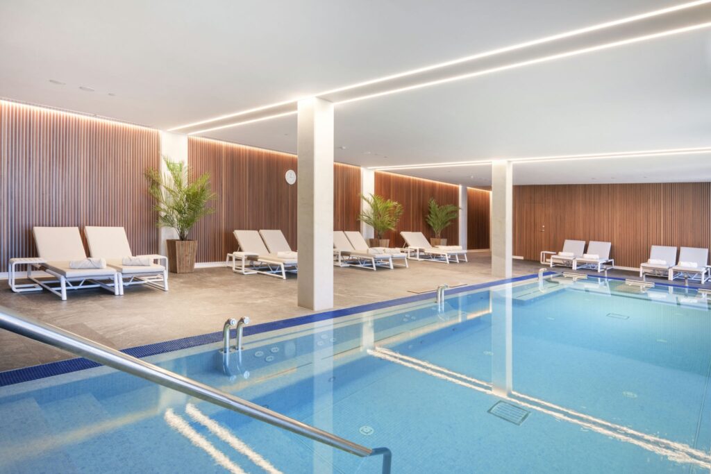 Serena Área de Spa Interior con Piscina Clara y Vegetación Exuberante Moderno spa interior con una piscina clara con barandilla de acero inoxidable, rodeada de cómodas tumbonas blancas y exuberantes plantas verdes, acentuado con elegantes paneles de madera y cálida iluminación ambiental.