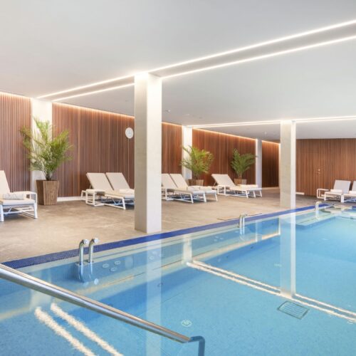 Moderno spa interior con una piscina clara con barandilla de acero inoxidable, rodeada de cómodas tumbonas blancas y exuberantes plantas verdes, acentuado con elegantes paneles de madera y cálida iluminación ambiental.