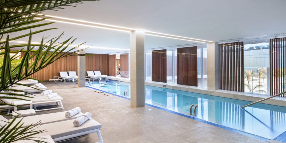 Un sereno área de piscina interior diseñada con líneas elegantes y una estética moderna, que cuenta con cómodas tumbonas adornadas con toallas enrolladas, una piscina clara y tranquila, y amplias ventanas que permiten que la abundante luz natural inunde el espacio. Plantas decorativas exuberantes añaden un toque de naturaleza, realzando la atmósfera tranquila y relajante similar a un spa, ideal para la rejuvenecimiento y el ocio.