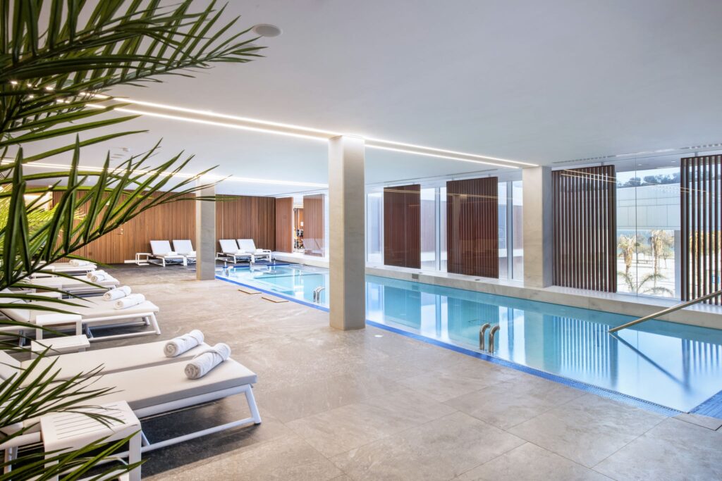 Moderna Piscina Interior con Diseño Elegante y Luz Natural Un sereno área de piscina interior diseñada con líneas elegantes y una estética moderna, que cuenta con cómodas tumbonas adornadas con toallas enrolladas, una piscina clara y tranquila, y amplias ventanas que permiten que la abundante luz natural inunde el espacio. Plantas decorativas exuberantes añaden un toque de naturaleza, realzando la atmósfera tranquila y relajante similar a un spa, ideal para la rejuvenecimiento y el ocio.
