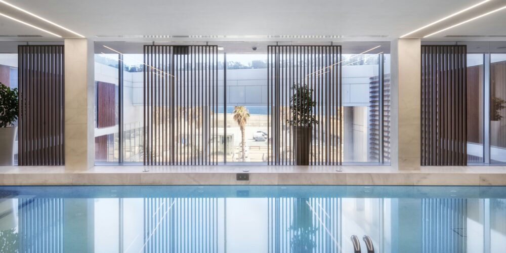 Una serena piscina interior cuenta con grandes ventanales que dejan entrar la luz natural, reflejando una arquitectura moderna con elegantes persianas verticales y vegetación. El diseño crea una atmósfera relajante perfecta para retiros de bienestar.
