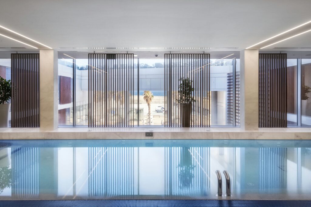 Tranquila piscina interior rodeada de luz natural Una serena piscina interior cuenta con grandes ventanales que dejan entrar la luz natural, reflejando una arquitectura moderna con elegantes persianas verticales y vegetación. El diseño crea una atmósfera relajante perfecta para retiros de bienestar.