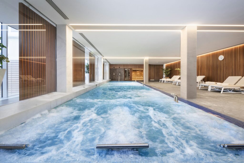 Tranquila piscina cubierta con arquitectura moderna y luz natural Una serena área de piscina interior que presenta una superficie de agua azul calmante con suaves olas, luces de techo modernas, grandes ventanas que permiten la entrada de luz natural, superficies blancas elegantes, acentos de madera cálida y sillas de lounge organizadas de manera ordenada para la relajación.