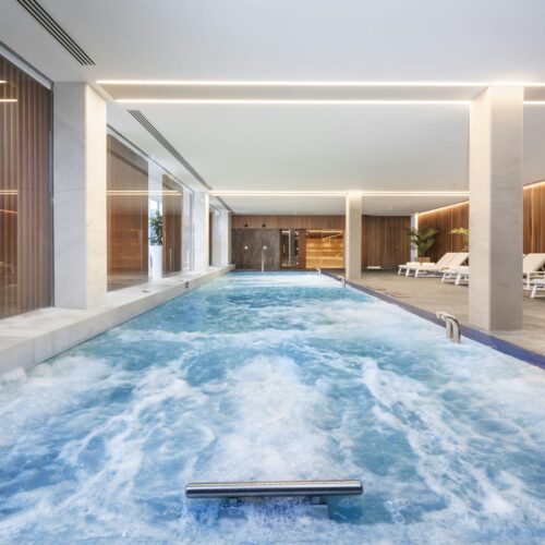 Una serena área de piscina interior que presenta una superficie de agua azul calmante con suaves olas, luces de techo modernas, grandes ventanas que permiten la entrada de luz natural, superficies blancas elegantes, acentos de madera cálida y sillas de lounge organizadas de manera ordenada para la relajación.