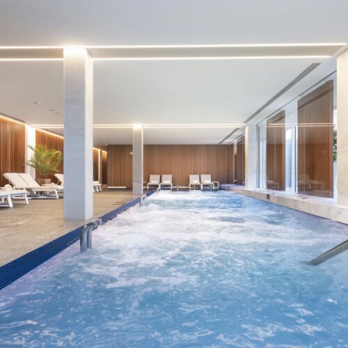 Área moderna de piscina cubierta que presenta una vibrante piscina azul con suaves olas, elegantes tumbonas blancas sobre un suelo de piedra clara, paneles de madera en las paredes y abundante luz natural a través de grandes ventanales de vidrio.