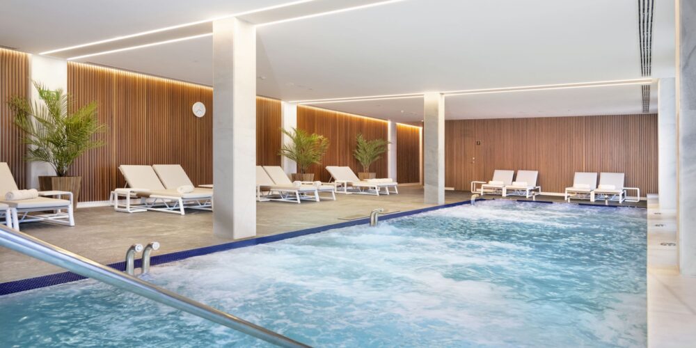 Spa interior moderno que cuenta con una piscina relajante con agua burbujeante, sillas de salón blancas y plantas en macetas verdes, complementado por paneles de madera cálida y una iluminación suave para una atmósfera tranquila.