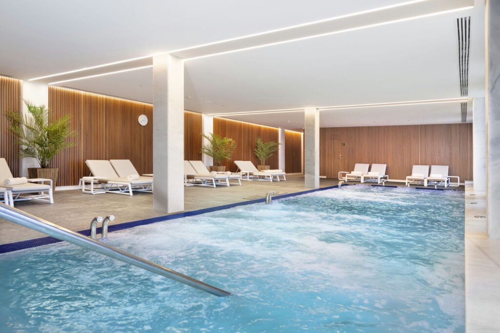 Área de spa interior serena con piscina burbujeante y salón de relajación Spa interior moderno que cuenta con una piscina relajante con agua burbujeante, sillas de salón blancas y plantas en macetas verdes, complementado por paneles de madera cálida y una iluminación suave para una atmósfera tranquila.